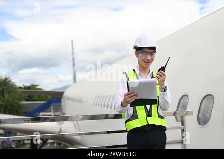 Junger asiatischer Mann in einer gelben Weste mit Walkie-Talkie, der auf einem Metallgeländer neben einem Flugzeug steht Stockfoto