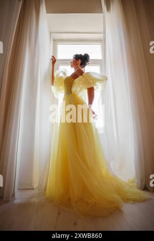 Die Silhouette einer Frau in einem goldenen, luxuriösen Kleid vor dem Hintergrund eines Fensters hält einen Vorhang mit den Händen. Elegante Dame in gelber Seide Stockfoto