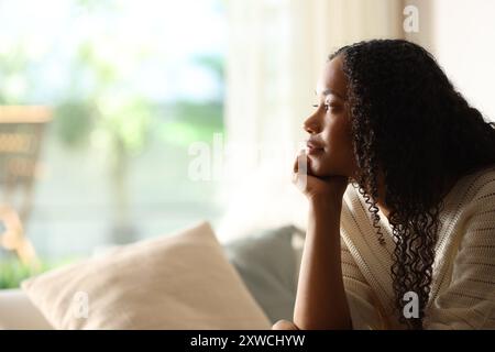 Schwarze Frau, die durch ein Fenster nachdenkt und zu Hause auf einer Couch sitzt Stockfoto