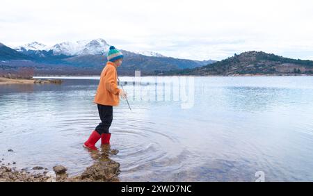 Kaukasischer Junge mit warmen Kleidern und Gummistiefeln, der in einem See mit verschneiten Bergen im Hintergrund spielt und Kopierraum. Konzeptspiele im Freien Stockfoto