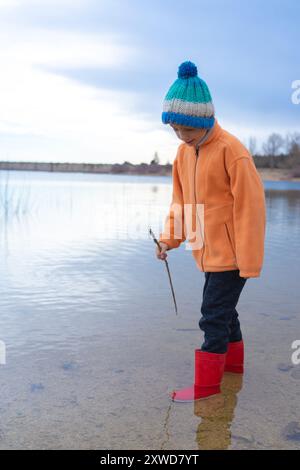 Ein kaukasischer Junge, der in einem See in der Natur spielt, trägt Gummistiefel, einen orangefarbenen Mantel und einen Wollhut mit Kopierraum Stockfoto