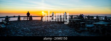 Eine Gruppe von Einzelpersonen steht an einem Aussichtspunkt und genießt den atemberaubenden Blick auf den Sonnenuntergang. Warme Farbtöne bemalen den Himmel, während die Sonne untergeht, und schaffen einen perfekten Moment für Reflexion und Verbindung. Stockfoto