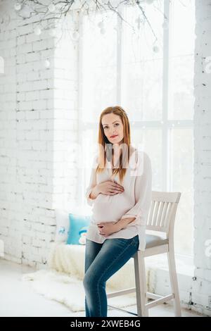 Ein rothaariges schwangeres Mädchen in einer hellen Bluse und einer blauen Jeans sitzt auf einem Hochstuhl in einem hellen Raum Stockfoto