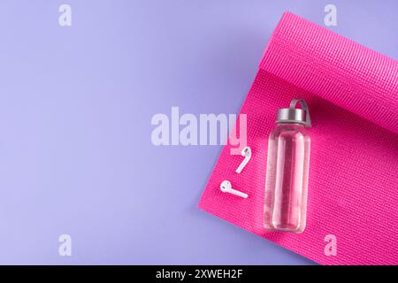 Sport- und Fitness-Hintergrund. Sportaccessoires für Damen. Yogamatte mit Kurzhanteln, Flasche Wasser, Handtuch, Sprungseil, Kopfhörer, auf hellvioletter Rückseite Stockfoto