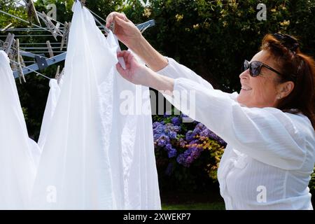Reife Frau, die weiße Bettlaken an einer rotierenden Wäscheleine hängt - John Gollop Stockfoto