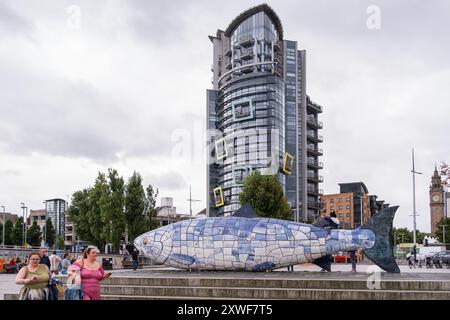 Belfast, Vereinigtes Königreich - 17. August 2024: Ufer des Lagan River mit Blick auf die Albert-Uhr in der Ferne. Im Vordergrund, zwei Frauen, die an der Skulptur Big Fish vorbeikommen. Stockfoto