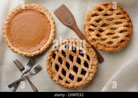 Hochwinkelaufnahme von drei frisch gebackenen hausgemachten Kuchen, Apfel, Kirsche und Kürbis auf einem Sackleinen-Tischtuch. Horizontales Format mit Gabeln und Holzspachtel Stockfoto