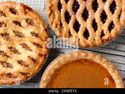 Drei Kuchen auf Kühlregalen. Großer Nahaufnahme von frisch gebackenen Apfel-, Kirsch- und Kürbiskuchen auf Drahtgestellen auf einem rustikalen Küchentisch aus Holz. Horiz Stockfoto