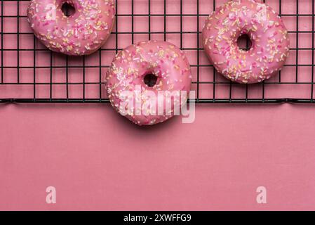 Drei rosafarbene Donuts mit bunten Streuseln ruhen teilweise auf einem Kühlregal auf einem rosafarbenen Hintergrund und strahlen ein einladendes und köstliches aus Stockfoto
