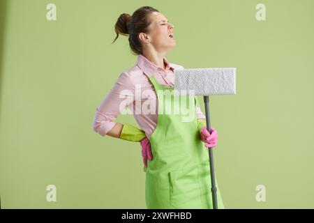 Frühjahrsputz. Traurige moderne 40-jährige Frau in grüner Schürze und Gummihandschuhen vor grünem Hintergrund mit Mopp und Rückenschmerzen. Stockfoto