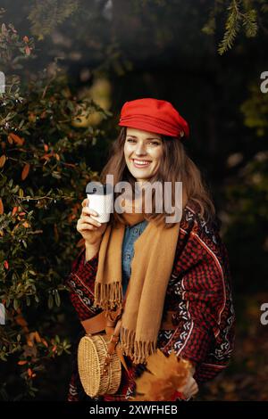 Hallo Herbst. Glückliche elegante 40-jährige Frau mit rotem Hut mit Herbstblättern, Schal, Tasche und Kaffee im Stadtpark. Stockfoto