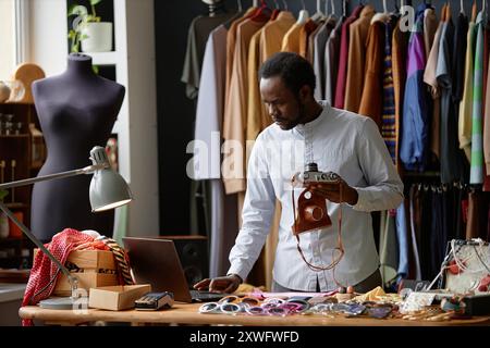Männlicher afroamerikanischer Second-Hand-Ladenassistent in elegantem Hemd mit Laptop, der eine alte Fotokamera zum Verkauf bringt, während er in sozialen Netzwerken im Second-Hand-Shop, Kopierraum, aufnimmt Stockfoto