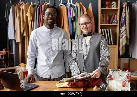 Mittleres Porträt von zwei glücklichen Second Hand Shop Assistenten in Vintage Brille lächeln, während sie die Kamera an der Kasse eines Second Hand Shops betrachten Stockfoto