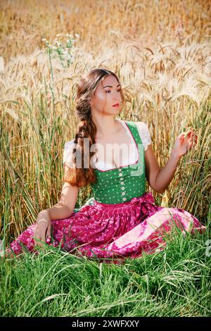 Eine junge Frau sitzt auf Einem weiten Maisfeld und trägt Ein traditionelles bayerisches Dirndl. Ihr Langes, Braunes Haar Ist Locker Über Ihre Schultern Und Sie Radiat Stockfoto