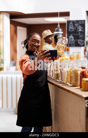 Black Shopkeeper wirbt für Bio-Produkte und bindet Kunden über soziale Medien und Videoanrufe in Bio-Food-Geschäften. Sie präsentiert eine Vielzahl frischer Artikel in Glasgläsern und Verpackungen ohne Abfall. Stockfoto