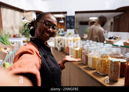 POV eines afroamerikanischen Öko-Ladeninhabers, der nachhaltige Lebensmittel fördert. Gäste kaufen frische, nahrhafte Speisen in einem modernen Supermarkt ein. Online-Videoanrufe zeigen umweltfreundliche Produkte. Stockfoto