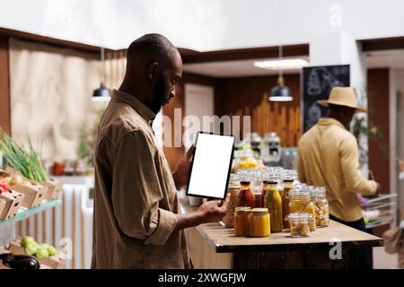 Ein afroamerikanischer Mann überprüft die umweltfreundlichen Produkte, Gläser und Lebensmittel in Regalen, während er ein Tablet mit einer Chromakey-Vorlage hält. Leere Bildschirme für Modelle. Nachhaltiger lokaler Supermarkt. Stockfoto
