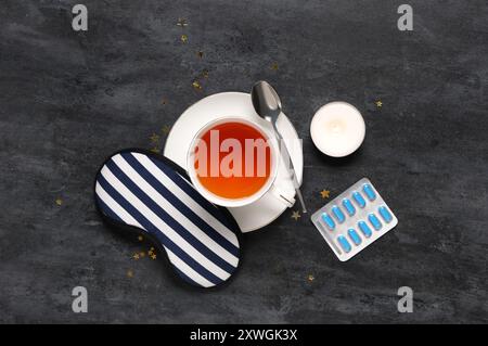 Tasse Tee mit Schlafmaske, Pillen und brennender Kerze auf schwarzem Hintergrund Stockfoto