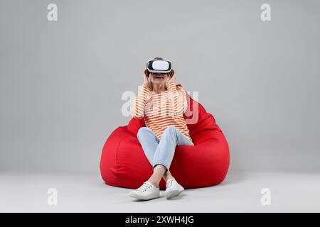 Glückliche Frau mit Virtual-Reality-Headset auf Sitzsack-Stuhl vor grauem Hintergrund Stockfoto
