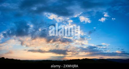 Atemberaubender Himmel voller dramatischer Wolken übergeht bei Sonnenuntergang von tiefem Blau zu sanften Pastelltönen. Horizont gesäumt von silbrigen Hügeln und Wäldern, die der ruhigen und weitläufigen Landschaft Tiefe verleihen. Stockfoto