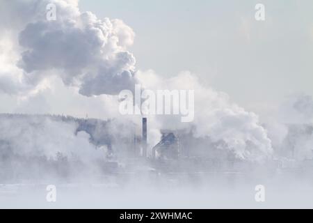 An einem frostigen Tag steigt Rauch aus mehreren Fabrikschornsteinen auf. Nebel breitet sich im Vordergrund aus. Stockfoto