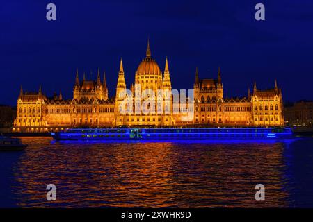 Budapest, Ungarn - 7. Juli 2024: Beleuchtetes parlamentsgebäude an der Donau bei Nacht mit einem hell beleuchteten Boot im Vordergrund, das eine Betäubung erzeugt Stockfoto