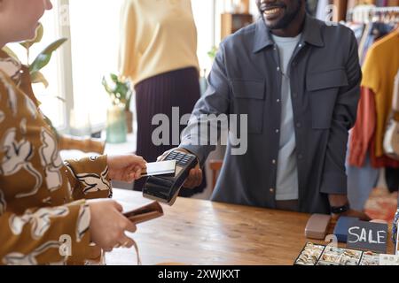 Zugeschnittene Aufnahme eines unmerklich lächelnden afroamerikanischen Kassierers mit POS-Terminal, während weibliche Kunden im Second-Hand-Geschäft drahtlos bezahlen, Schwerpunkt auf Kartenleser, Kopierraum Stockfoto