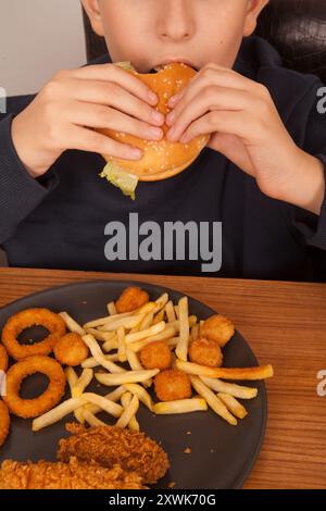 Ein Kaukasier isst halbgefressenen Hamburger, Kartoffelchips, Zwiebelringe und Hühnchenstücke von Hand von einem grauen Teller auf einem Holztisch, amerikanischer schneller Foo Stockfoto