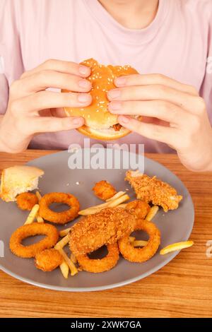 Ein Kaukasier isst halbgefressenen Hamburger, Kartoffelchips, Zwiebelringe und Hühnchenstücke von Hand von einem grauen Teller auf einem Holztisch, amerikanischer schneller Foo Stockfoto