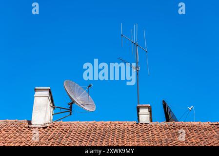 TV-Antennen im alten Stil und Satellitenschüssel auf dem Dach des Gebäudes mit roten Fliesen Stockfoto