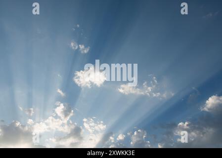 Sonnenstrahlen, die von hinter Wolken über einen blauen Himmel schweben, erhellen den Dunst der hohen Atmosphäre Stockfoto