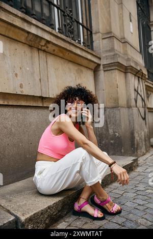 Eine Frau in rosa-weißem Outfit sitzt auf einer Stufe in einer europäischen Stadt und spricht mit ihrem Telefon. Stockfoto