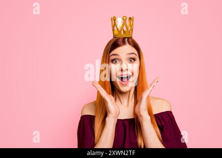 Eine Frau mit dem überraschten Gesicht und der Krone auf dem Kopf öffnete den Mund und die Augen weit und hielt die Hände in der Nähe ihrer Wangen, isoliert auf gelbem Hintergrund Stockfoto