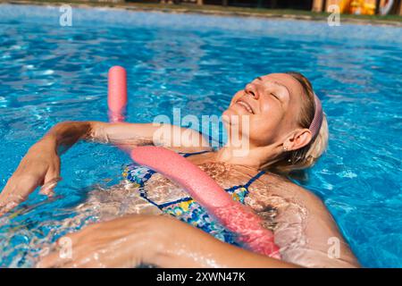 Eine Seniorin genießt einen sonnigen Tag im Pool mit einer rosa Nudel. Sie wirkt zufrieden und entspannt und saugt die Wärme auf Stockfoto