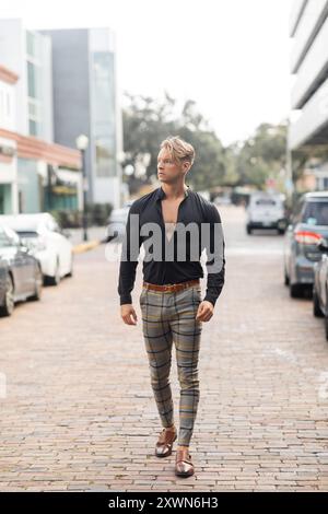 Ein gutaussehender blonder Mann läuft selbstbewusst eine Straße hinunter. Er trägt ein stylisches schwarzes Hemd und karierte Hosen. Stockfoto