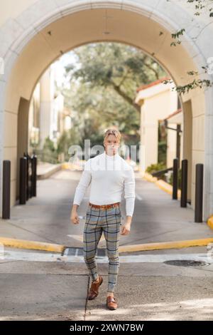 Ein blonder Mann mit weißem Rollkragen und karierter Hose posiert auf einer Straße in Florida. Stockfoto