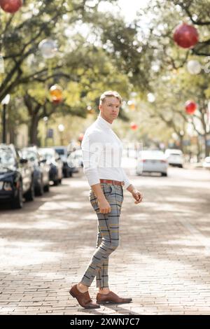 Ein blonder Mann in einem weißen Hemd und karierten Hosen posiert auf einer von Bäumen gesäumten Straße Floridas. Stockfoto