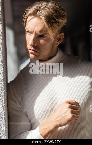 Ein blonder Mann in einem weißen Rollkragen steht vor einem Gebäude in Florida und sonnt sich im Sonnenlicht. Stockfoto