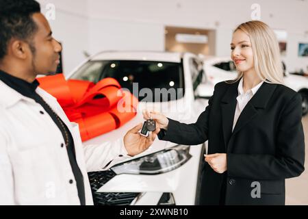 Nahaufnahme eines glücklichen afrikanischen Autobesitzers, der den Schlüssel von einer lächelnden Autohändlerin im Händlerbetrieb bekommt. Fröhlicher schwarzer Mann, der den Schlüssel zum gekauften Auto nimmt und versiegelt Stockfoto