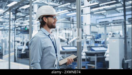 Junger Elektronikentwickler lächelnd, der auf die Produktionslinie blickt. Porträt eines Mannes, der einen weißen Schutzhelm trägt. Spezialist für die Überwachung künstlicher Intelligenzsysteme in einer Fabrik Stockfoto