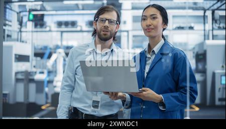 Industrieingenieure, die in einer Technologiefabrik arbeiten. Kaukasischer Mann und asiatische Frau mit Laptop-Computer und Unterhaltung. Filmmaterial mit einem Porträt von ein paar Mitarbeitern in einem Werk Stockfoto