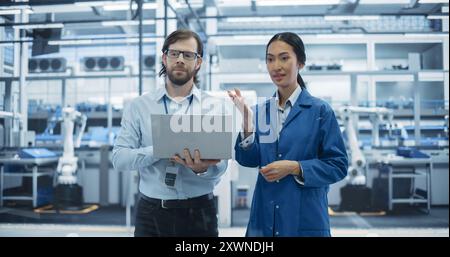 Kaukasischer Mann und asiatische Frau haben ein Gespräch in einer Industrieanlage. Fortgeschrittene Fabrikmitarbeiter arbeiten zusammen und nutzen Laptop-Computer zur Überwachung der Arbeitsprozesse in einer modernen Anlage Stockfoto
