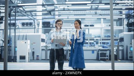Kaukasischer Mann und asiatische Frau, die sich in einer Industrieanlage unterhalten. Fortgeschrittene Werksmitarbeiter arbeiten zusammen und nutzen Laptop-Computer zur Überwachung des Arbeitsprozesses in einer Anlage Stockfoto
