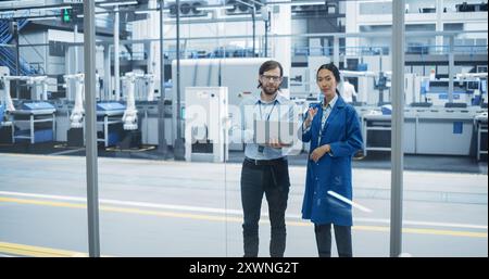 Kaukasischer Mann und asiatische Frau, die sich in einer Industrieanlage unterhalten. Fortgeschrittene Fabrikmitarbeiter arbeiten zusammen und nutzen Laptop-Computer zur Überwachung der Arbeitsprozesse in einer modernen Anlage Stockfoto