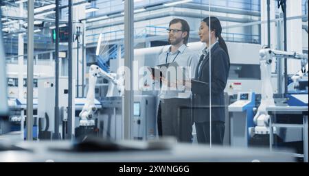 Techniker und Geschäftsfrau stehen im Büro, sprechen, verwenden Computer und überwachen die Produktion in einer modernen automatisierten Elektronikfertigung mit KI Roboterarmen Stockfoto