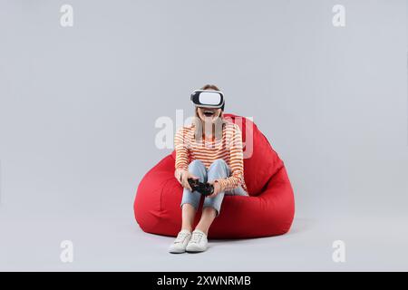 Glückliche Frau mit Virtual-Reality-Headset und Controller auf Sitzsack-Stuhl vor grauem Hintergrund Stockfoto