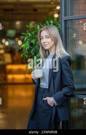 Lächelnde, selbstbewusste Geschäftsfrau trinkt Kaffee während der Pause, mit Blick auf die Kamera am Eingang des Cafés im Freien Stockfoto