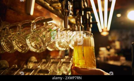 Der Barkeeper hält ein Glas Lagerbier, das aus dem Zapfhahn gegossen wird, wobei Reihen leerer Gläser über und hinter dem Zapfhahn hängen. Stockfoto