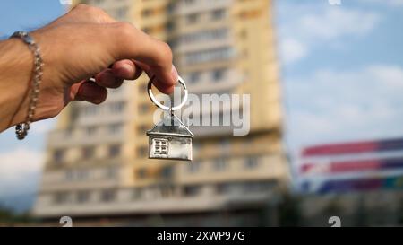 Hand hält einen Schlüsselanhänger in Form eines Hauses gegen verschwommenes Wohngebäude. Immobilien, Eigentum, neues Hauskonzept. Stockfoto