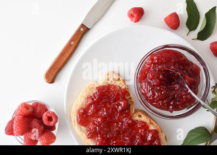 Frühstück mit Himbeermarmelade in einer Glasschale auf einem weißen Tisch mit einer Schüssel voller Beeren und Brotscheiben. Draufsicht. Stockfoto